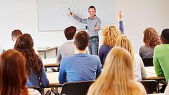Lehrer steht vor der Klasse an einem Whiteboard und erklärt etwas