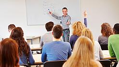Lehrer steht vor der Klasse an einem Whiteboard und erklärt etwas