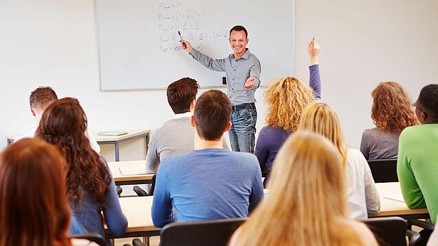 Lehrer steht vor der Klasse an einem Whiteboard und erklärt etwas