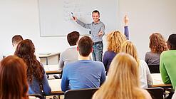 Lehrer steht vor der Klasse an einem Whiteboard und erklärt etwas