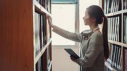 Eine junge Frau greift nach Büchern in einem Regal.