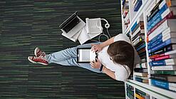 Eine Studentin sitzt in einer Bibliothek auf dem Gang und tippt auf einem Tablet.