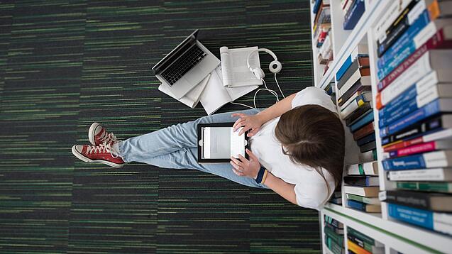 Eine Studentin sitzt in einer Bibliothek auf dem Gang und tippt auf einem Tablet.