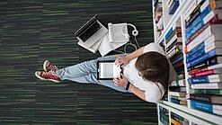 Eine Studentin sitzt in einer Bibliothek auf dem Gang und tippt auf einem Tablet.