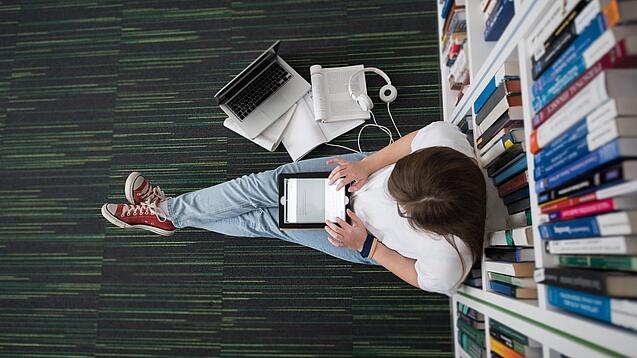 Eine Studentin sitzt in einer Bibliothek auf dem Gang und tippt auf einem Tablet.