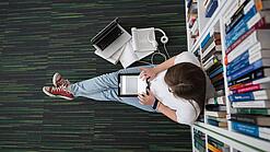 Eine Studentin sitzt in einer Bibliothek auf dem Gang und tippt auf einem Tablet.
