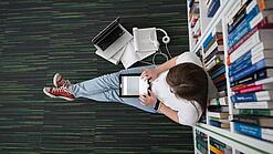 Eine Studentin sitzt in einer Bibliothek auf dem Gang und tippt auf einem Tablet.