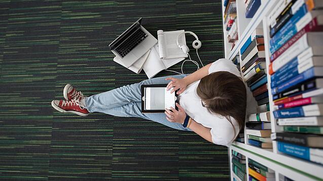 Eine Studentin sitzt in einer Bibliothek auf dem Gang und tippt auf einem Tablet.