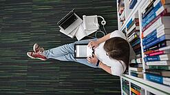 Eine Studentin sitzt in einer Bibliothek auf dem Gang und tippt auf einem Tablet.