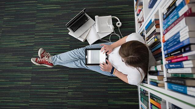 Eine Studentin sitzt in einer Bibliothek auf dem Gang und tippt auf einem Tablet.
