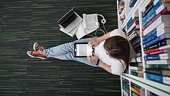 Eine Studentin sitzt in einer Bibliothek auf dem Gang und tippt auf einem Tablet.