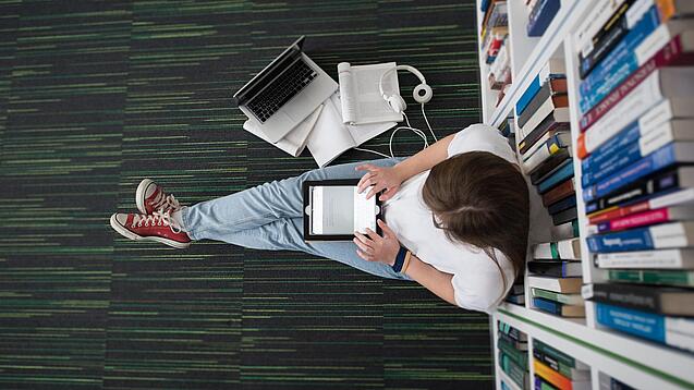 Eine Studentin sitzt in einer Bibliothek auf dem Gang und tippt auf einem Tablet.
