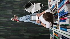 Eine Studentin sitzt in einer Bibliothek auf dem Gang und tippt auf einem Tablet.