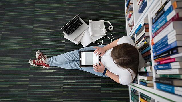 Eine Studentin sitzt in einer Bibliothek auf dem Gang und tippt auf einem Tablet.