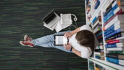 Eine Studentin sitzt in einer Bibliothek auf dem Gang und tippt auf einem Tablet.