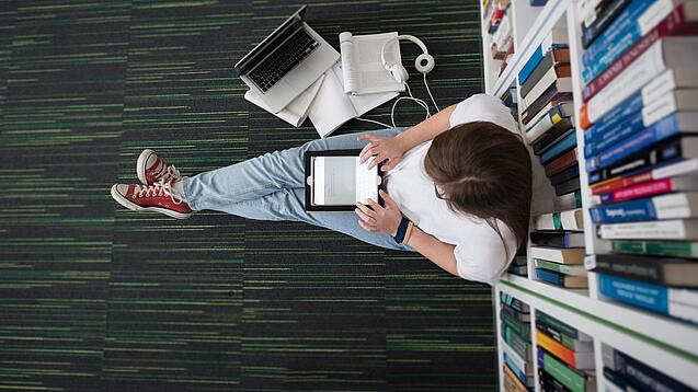 Eine Studentin sitzt in einer Bibliothek auf dem Gang und tippt auf einem Tablet.