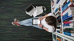 Eine Studentin sitzt in einer Bibliothek auf dem Gang und tippt auf einem Tablet.