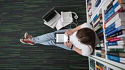 Eine Studentin sitzt in einer Bibliothek auf dem Gang und tippt auf einem Tablet.