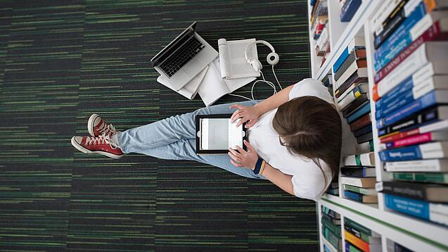 Eine Studentin sitzt in einer Bibliothek auf dem Gang und tippt auf einem Tablet.