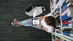 Eine Studentin sitzt in einer Bibliothek auf dem Gang und tippt auf einem Tablet.