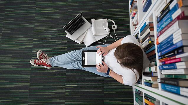 Eine Studentin sitzt in einer Bibliothek auf dem Gang und tippt auf einem Tablet.