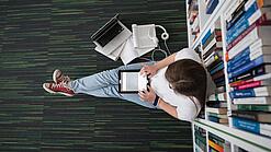 Eine Studentin sitzt in einer Bibliothek auf dem Gang und tippt auf einem Tablet.