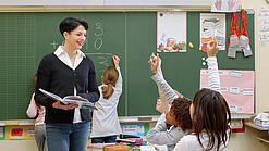 Eine Lehrerin steht vor einer Tafel, Schülerinnen melden sich.