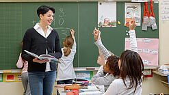 Eine Lehrerin steht vor einer Tafel, Schülerinnen melden sich.