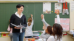Eine Lehrerin steht vor einer Tafel, Schülerinnen melden sich.