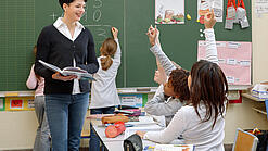 Eine Lehrerin steht vor einer Tafel, Schülerinnen melden sich.