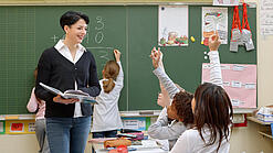 Eine Lehrerin steht vor einer Tafel, Schülerinnen melden sich.
