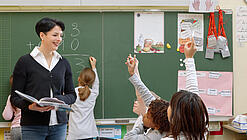 Eine Lehrerin steht vor einer Tafel, Schülerinnen melden sich.
