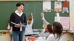 Eine Lehrerin steht vor einer Tafel, Schülerinnen melden sich.