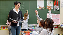 Eine Lehrerin steht vor einer Tafel, Schülerinnen melden sich.