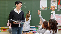 Eine Lehrerin steht vor einer Tafel, Schülerinnen melden sich.