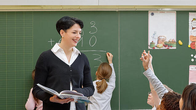 Eine Lehrerin steht vor einer Tafel, Schülerinnen melden sich.