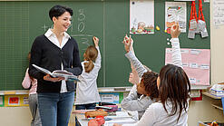 Eine Lehrerin steht vor einer Tafel, Schülerinnen melden sich.
