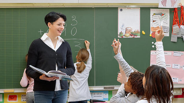 Eine Lehrerin steht vor einer Tafel, Schülerinnen melden sich.