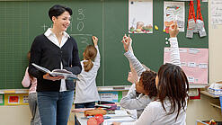 Eine Lehrerin steht vor einer Tafel, Schülerinnen melden sich.