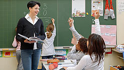 Eine Lehrerin steht vor einer Tafel, Schülerinnen melden sich.