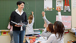 Eine Lehrerin steht vor einer Tafel, Schülerinnen melden sich.