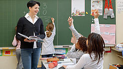 Eine Lehrerin steht vor einer Tafel, Schülerinnen melden sich.