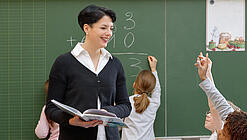 Eine Lehrerin steht vor einer Tafel, Schülerinnen melden sich.