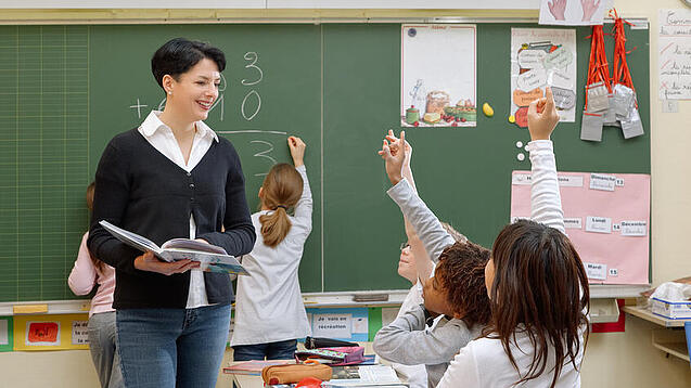 Eine Lehrerin steht vor einer Tafel, Schülerinnen melden sich.