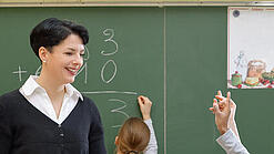 Eine Lehrerin steht vor einer Tafel, Schülerinnen melden sich.