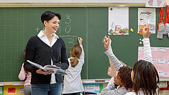 Eine Lehrerin steht vor einer Tafel, Schülerinnen melden sich.