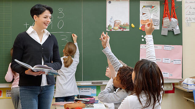 Eine Lehrerin steht vor einer Tafel, Schülerinnen melden sich.