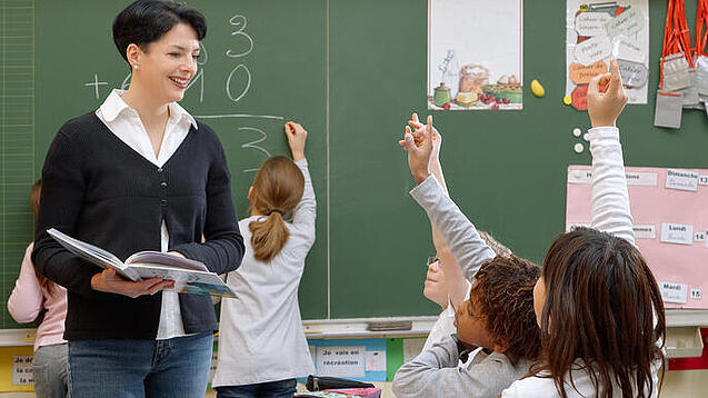 Eine Lehrerin steht vor einer Tafel, Schülerinnen melden sich.