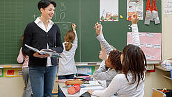 Eine Lehrerin steht vor einer Tafel, Schülerinnen melden sich.