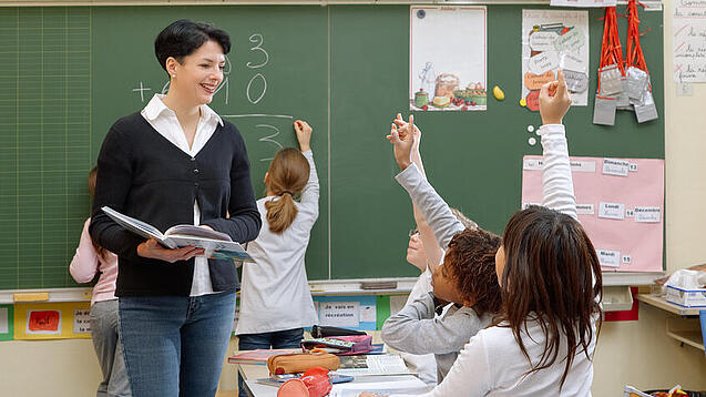 Eine Lehrerin steht vor einer Tafel, Schülerinnen melden sich.